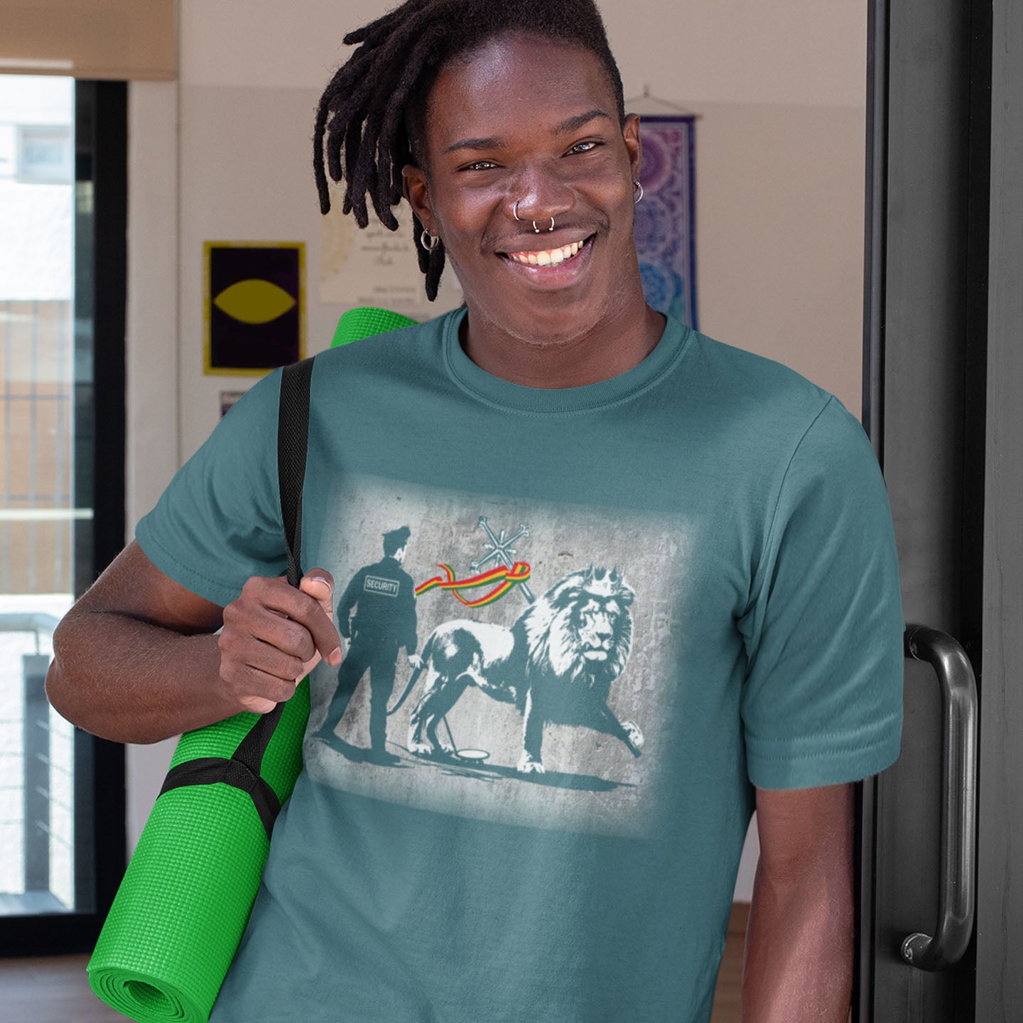 Person wearing a teal t-shirt with a graphic design, holding a green yoga mat. A Banksy style t-shirt in a stencil art mockery of how much security we have in the western world, from a Rastafarian view point. Made from 100% organic ring-spun cotton, this graffiti stencil art t-shirt is unisex, high-quality, super comfy, and best of all, eco-friendly.