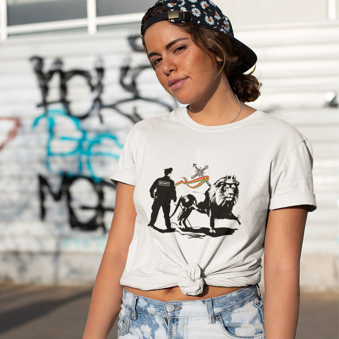 A woman wearing a baseball cap backwards standing in the street, wearing a white short sleeved crew neck t-shirt with a graffiti stencil design of a security guard checking the underside of the Lion of Judah for explosives.