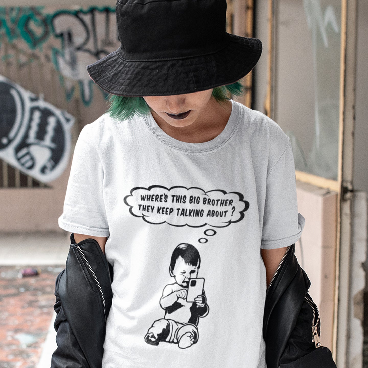 Person wearing a white t-shirt with a graphic and text, standing in an urban setting. A witty conspiracy theory t-shirt questioning the existence of a controlling 'Big Brother' watching over us. This unique design in stencil art illustrates the sweet mind of a child and a lesson in how to take things literally. Big Brother is everywhere and nowhere. Made from 100% organic ring-spun cotton, this Banksy style conspiracy theory t-shirt is unisex, high-quality, super comfy, and best of all, eco-friendly.