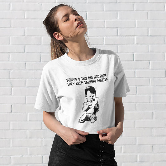 A woman wearing a white graphic t-shirt with a black cartoon baby and the text 'WHERE'S THIS BIG BROTHER THEY KEEP TALKING ABOUT?' posing against a white brick wall.