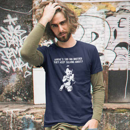 A man with long brown hair and a beard stands in front of a graffiti-covered wall, wearing a French navy blue graphic t-shirt over an olive green long-sleeve shirt. The t-shirt features a white graphic of a cartoon character and the text 'WHERE'S THIS BIG BROTHER THEY KEEP TALKING ABOUT?'.
