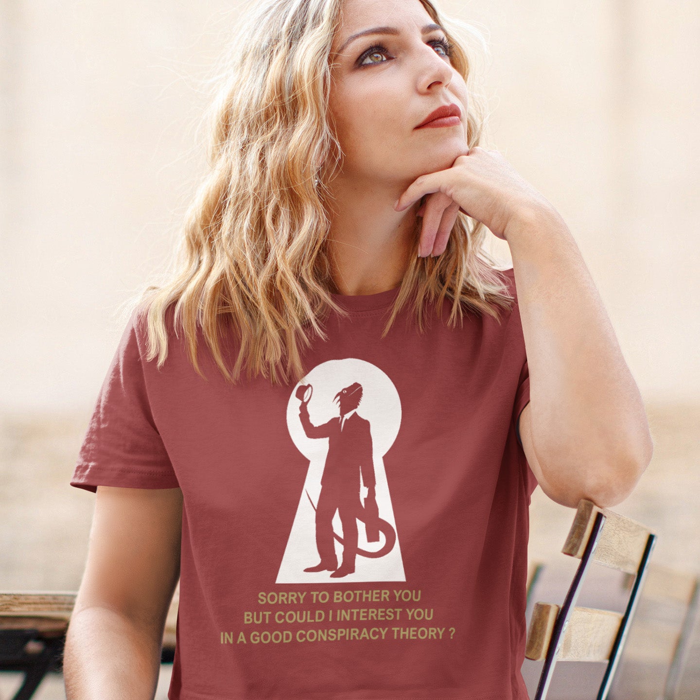 Woman wearing a maroon t-shirt with a graphic and text, sitting outdoors. A funny conspiracy theory t-shirt. Just an ordinary, everyday, door-to-door conspiracy theory salesman. Made from 100% organic ring-spun cotton, this reptilian alien t-shirt is unisex, high-quality, super comfy, and best of all, eco-friendly.