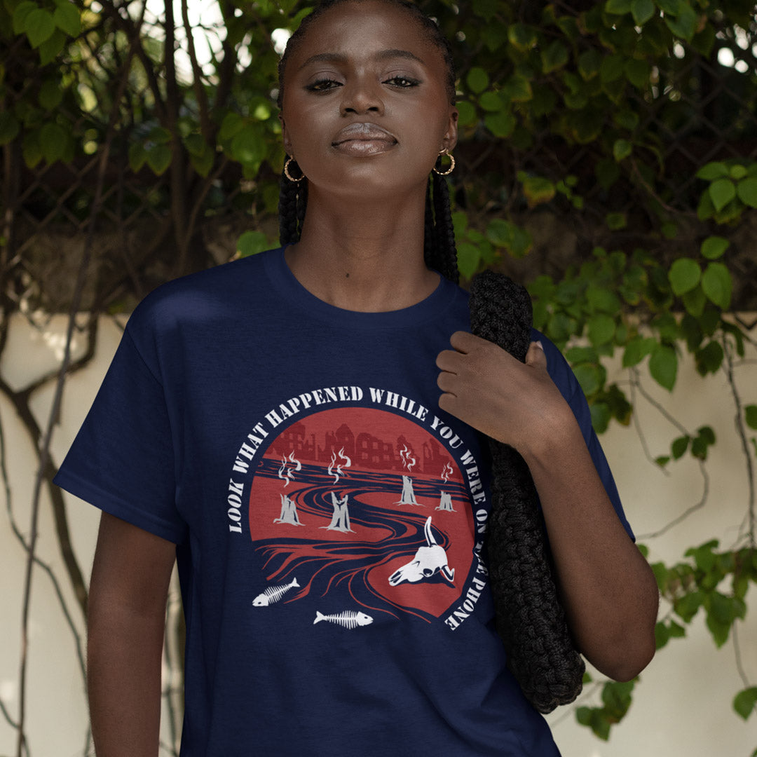 Woman wearing a navy blue t-shirt with a graphic design, standing outdoors with greenery in the background. An anti mobile phone abuse theme mixed with a save the earth t shirt. This design is a harsh view of what's happening to the planet while people are distracted by their phones. Made from 100% organic ring-spun cotton, this save the planet t shirt is unisex, high quality, super comfy, and best of all, eco-friendly.