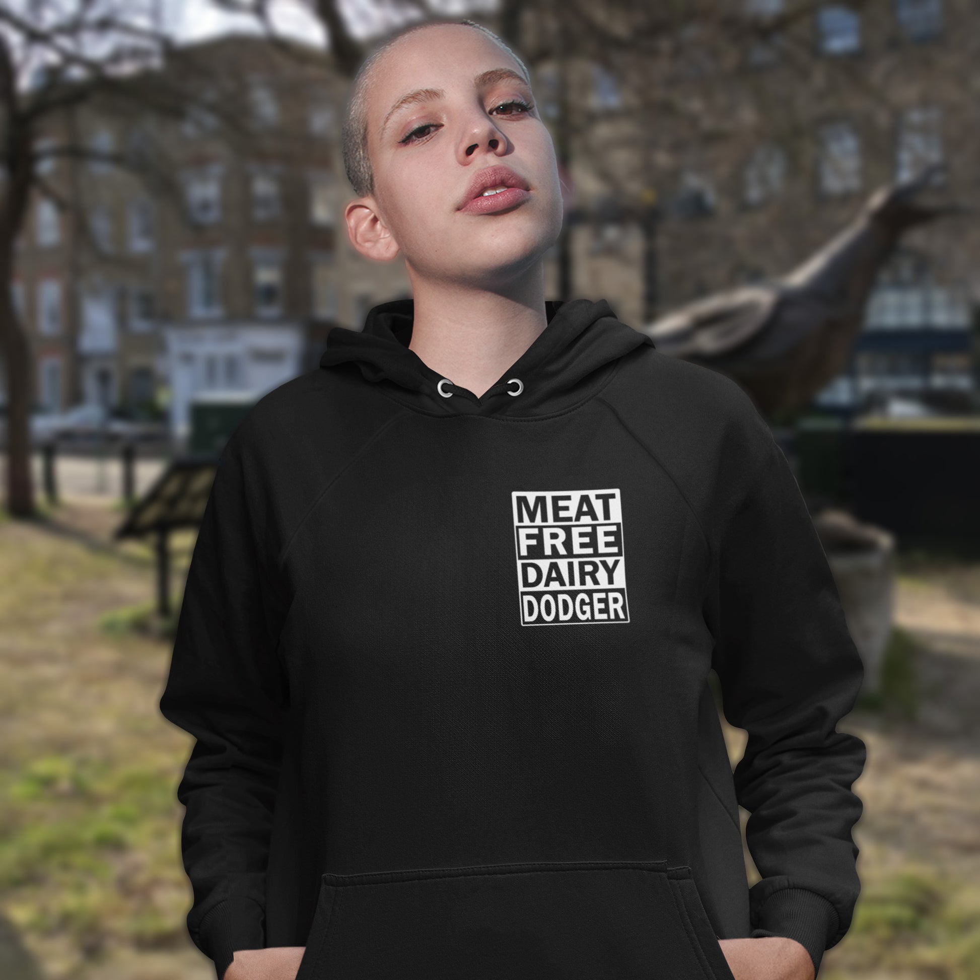 Person wearing a black hoodie with 'MEAT FREE DAIRY DODGER' text in an outdoor setting. This plant based vegan hoodie promotes a witty alternative to the 'non-dairy' label. Never mind 'plant based' as a new label for vegan . This is straight off the streets of East London. Comfortable and practical. It's extra soft and has a convenient front pouch pocket. Made from organic cotton and recycled polyester, this meat free hoodie design is a great eco-friendly choice.