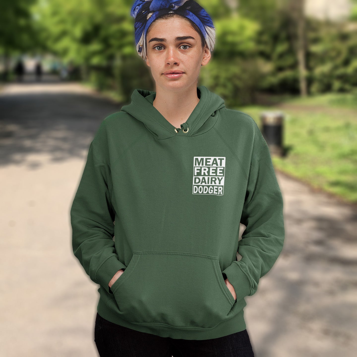 Person wearing a bottle green hoodie with 'MEAT FREE DAIRY DODGER' text in a park. This plant based vegan hoodie promotes a witty alternative to the 'non-dairy' label. Never mind 'plant based' as a new label for vegan . This is straight off the streets of East London. Comfortable and practical. It's extra soft and has a convenient front pouch pocket. Made from organic cotton and recycled polyester, this meat free hoodie design is a great eco-friendly choice.