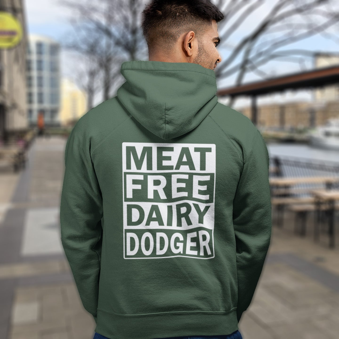 Person wearing a bottle green hoodie with 'MEAT FREE DAIRY DODGER' text on the back, standing outdoors. This vegan hoodie promotes a witty alternative to the 'non-dairy' label. Never mind 'plant based' as a new label for vegan . This is straight off the streets of East London. Comfortable and practical. It's extra soft and has a convenient front pouch pocket. Made from organic cotton and recycled polyester, this vegan hoodie is a great eco-friendly choice.