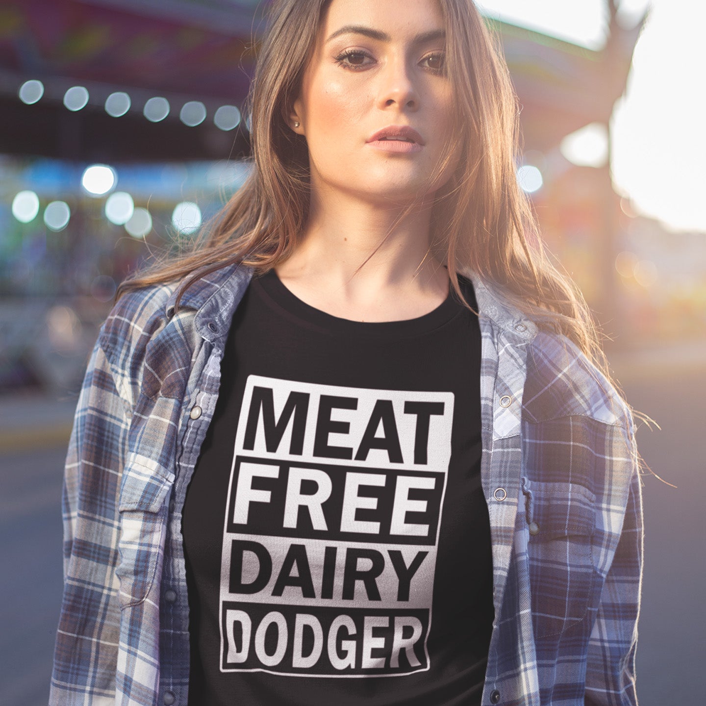 Woman wearing a black t-shirt with 'MEAT FREE DAIRY DODGER' text, standing in front of a blurred amusement park background. A vegan meat free t shirt promoting a witty alternative to the 'vegan' label. Never mind 'plant based' as a new label for vegan . This is straight off the streets of East London. Made from 100% organic ring-spun cotton, this vegan dairy free t shirt is unisex, high-quality, super comfy, and best of all, eco-friendly.