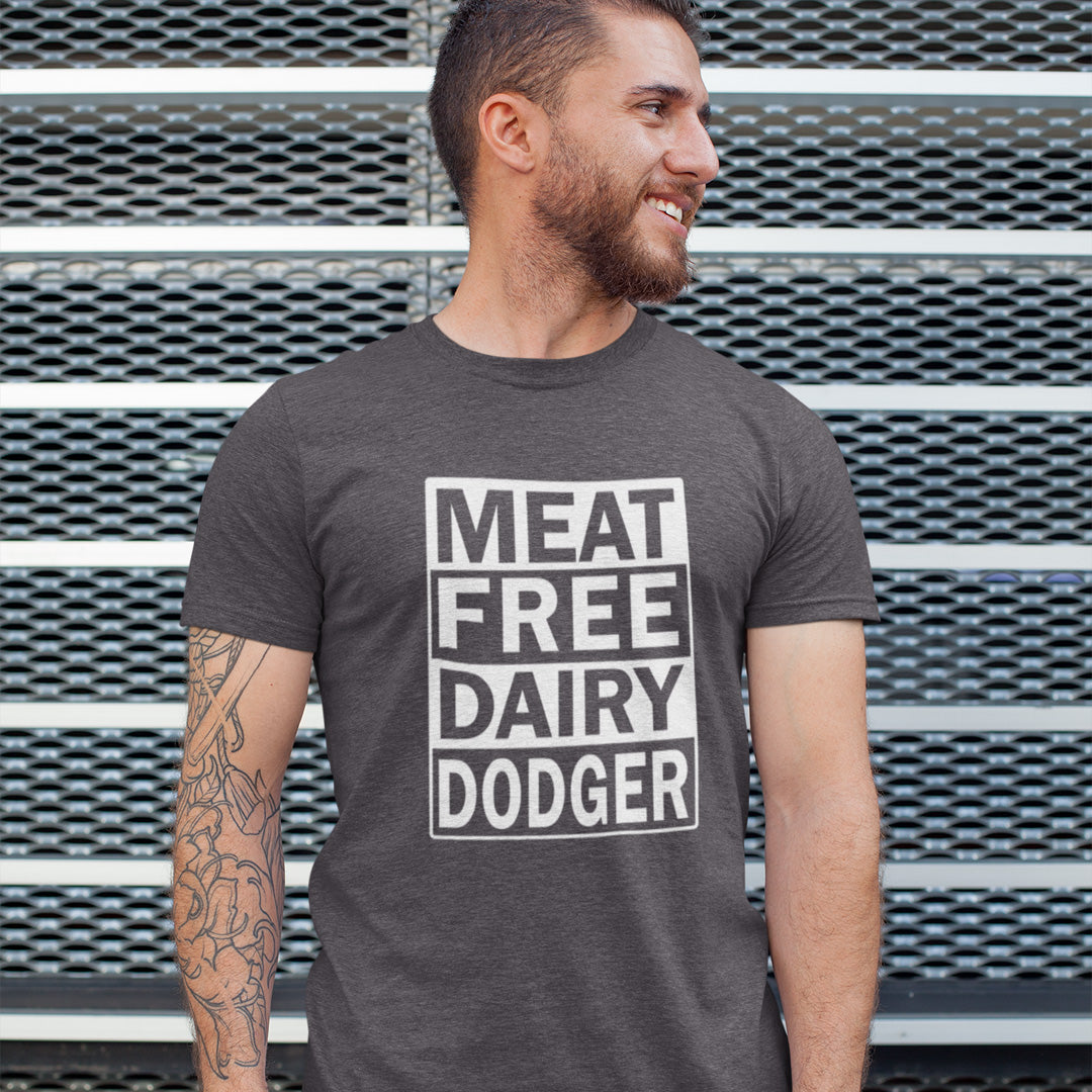 A man wearing an anthracite grey t-shirt with a white graphic that reads 'MEAT FREE DAIRY DODGER' in stacked text blocks, standing in front of a grey metal mesh wall.