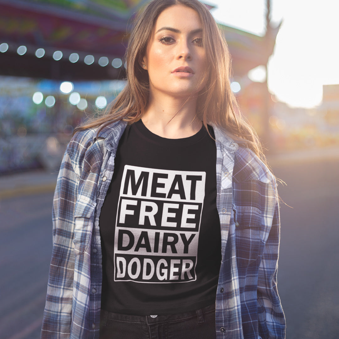 A woman wearing a black graphic t-shirt with the text 'MEAT FREE DAIRY DODGER' in bold white letters, styled with an open blue and white plaid flannel shirt over her shoulders, set against a blurred outdoor background.