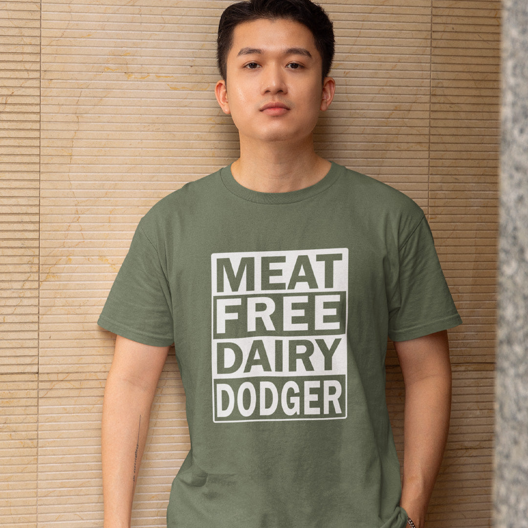 A man wearing an khaki green t-shirt with a white graphic box that reads 'MEAT FREE DAIRY DODGER', standing against a beige bamboo slat wall.