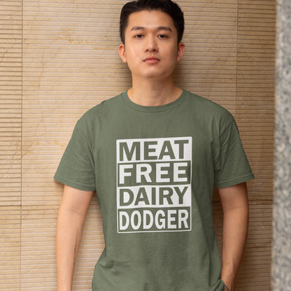 A man wearing an khaki green t-shirt with a white graphic box that reads 'MEAT FREE DAIRY DODGER', standing against a beige bamboo slat wall.