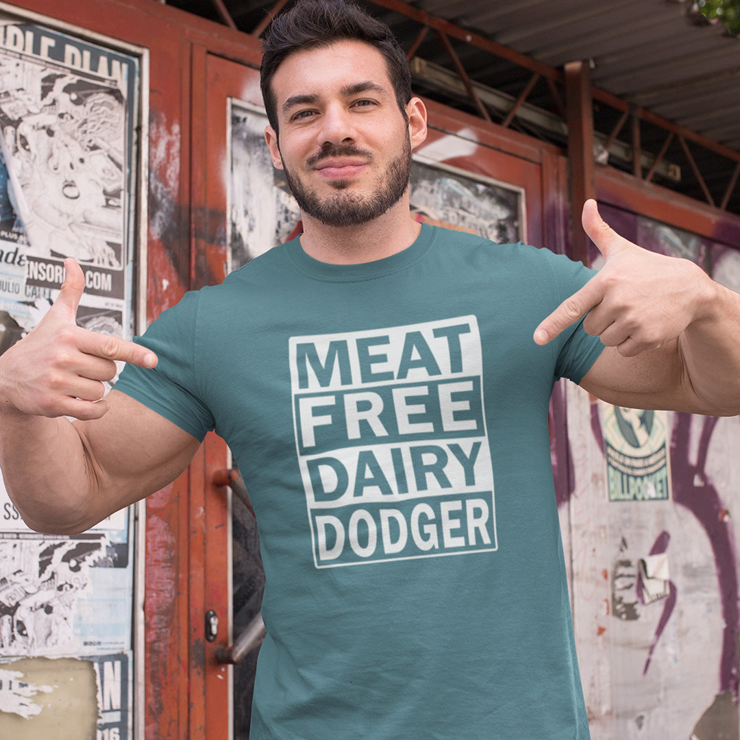 A man wearing a stargazer blue t-shirt with a white graphic that reads 'MEAT FREE DAIRY DODGER', posing with his thumbs pointing to the text in front of a graffiti-covered wall.