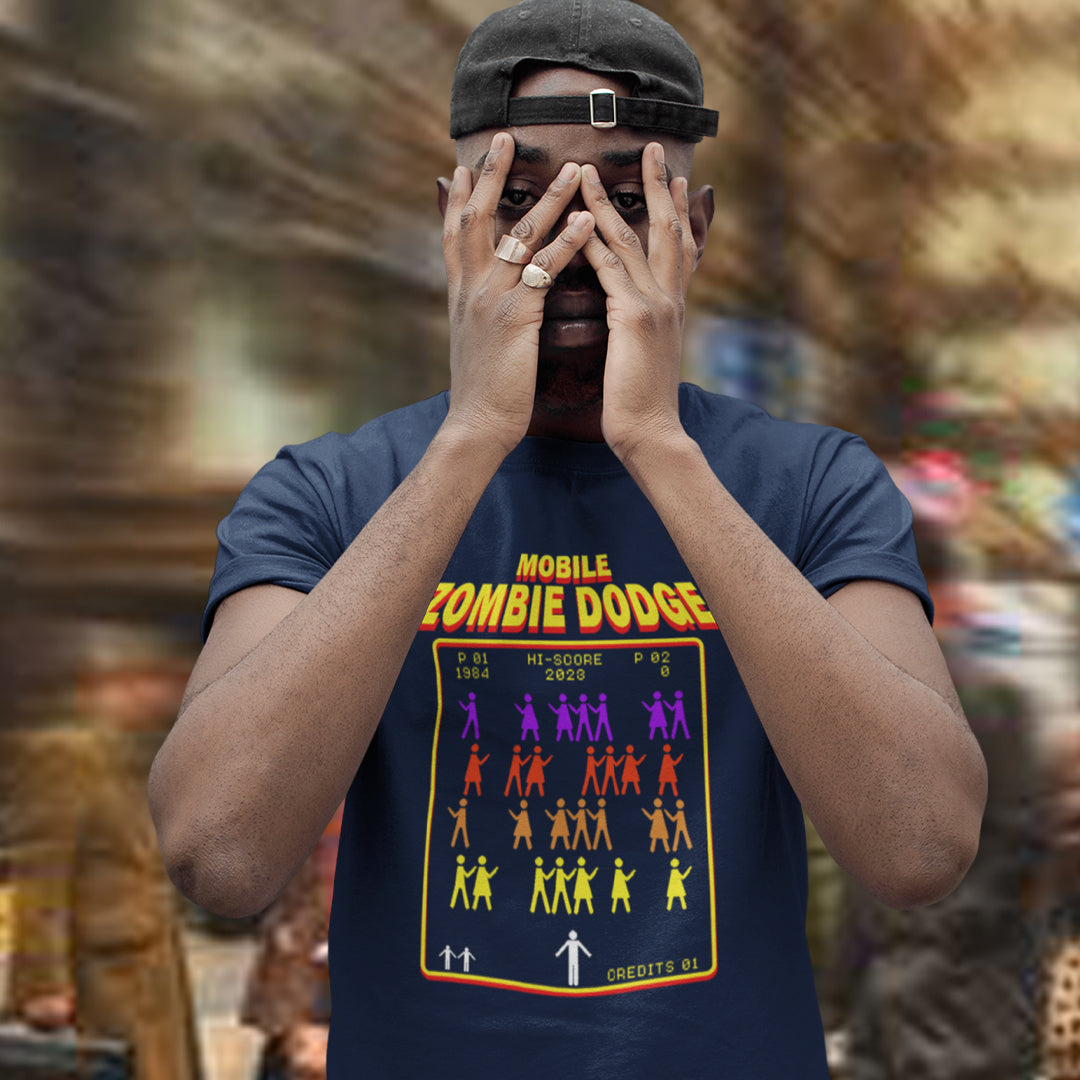 A man wearing a French navy blue graphic t-shirt with a retro arcade game design titled 'MOBILE ZOMBIE DODGE' and holding his hands over his eyes, set against a blurred outdoor background.