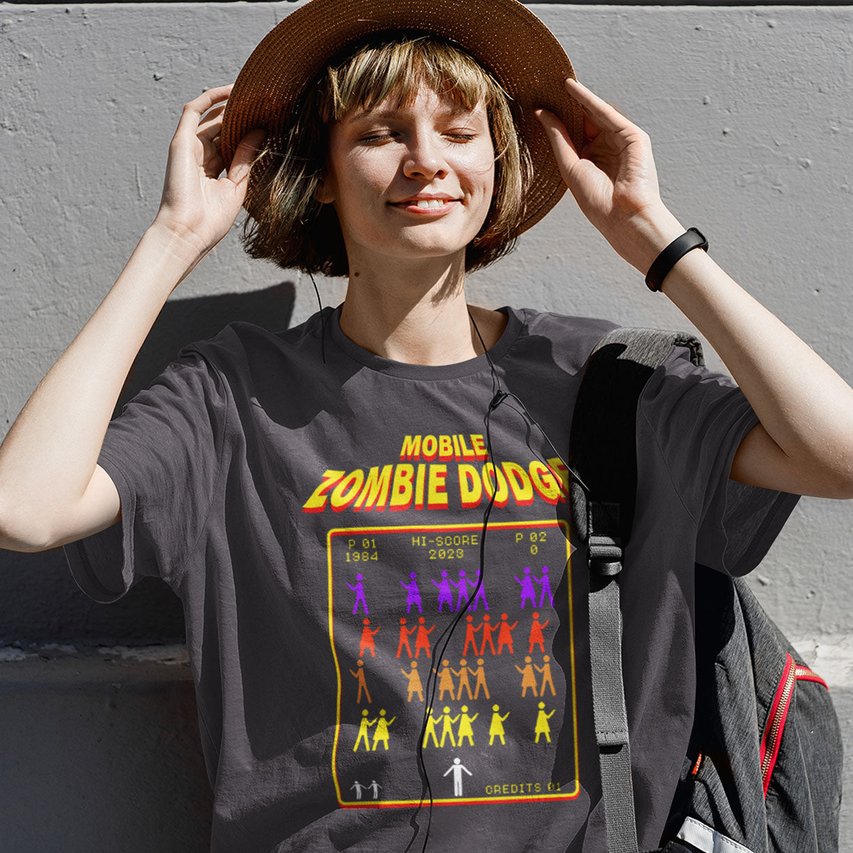 Person wearing a t-shirt with 'Mobile Zombie Dodge' graphic design against a grey wall. A satirical gamer t-shirt design for anyone who feels they are in a repetitive 80's video game every time they enter a crowded street scene. Welcome to the age of the mobile phone and lack of social awareness. Made from 100% organic ring-spun cotton, this unisex gamer t-shirt is alternative and original. It's high-quality, super comfy, and best of all—eco-friendly.