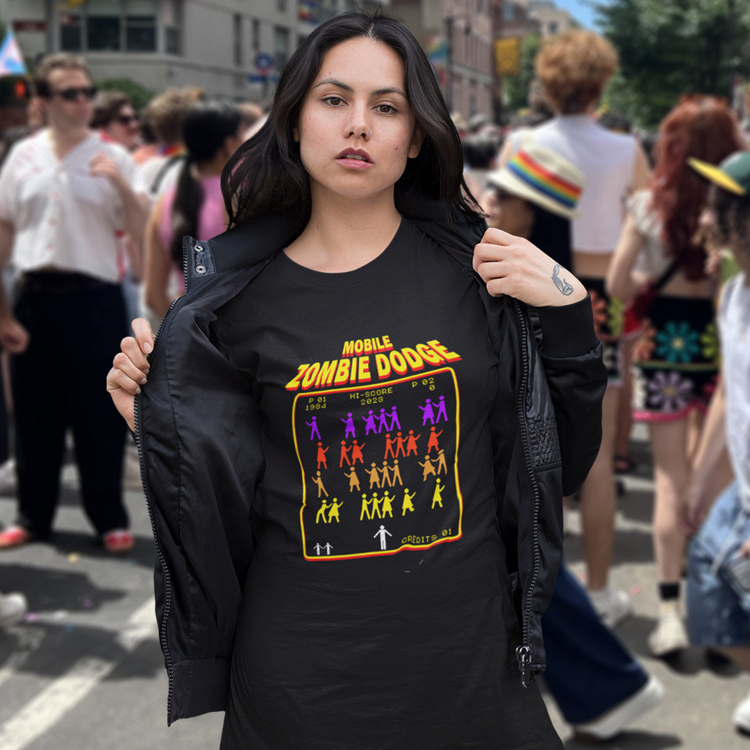 A woman standing outdoors at a street festival wearing a black graphic T-shirt featuring a retro video game style graphic on the chest. The graphic displays the text 'MOBILE ZOMBIE DODGE' in a yellow-orange gradient font above a grid of dancing zombie figures in purple, red, and yellow, mimicking a classic arcade game menu..