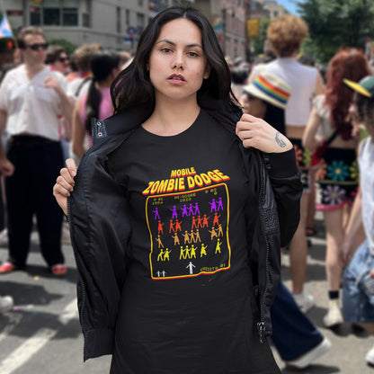 A woman standing outdoors at a street festival wearing a black graphic T-shirt featuring a retro video game style graphic on the chest. The graphic displays the text 'MOBILE ZOMBIE DODGE' in a yellow-orange gradient font above a grid of dancing zombie figures in purple, red, and yellow, mimicking a classic arcade game menu..