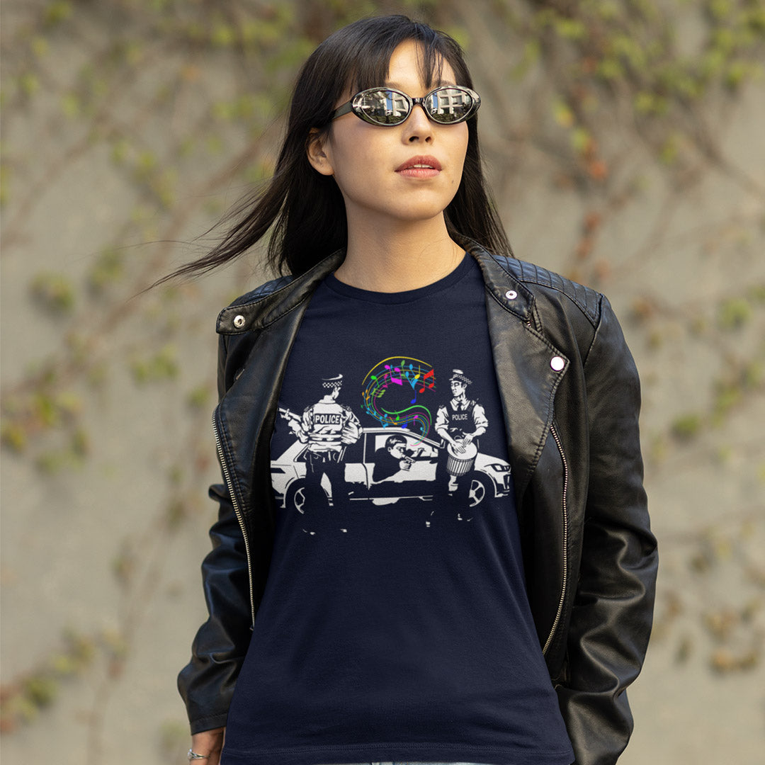 A woman wearing a French navy blue graphic t-shirt under a black leather jacket, styled with sunglasses, photographed outdoors against a blurred background of beige stone and green foliage.