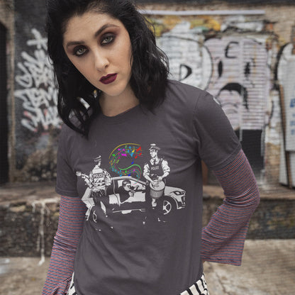 A woman wearing a dark gray graphic t-shirt with a black and white illustration of a band playing in front of a car, layered over a red and blue striped long-sleeve shirt. She is posing in front of a graffiti-covered wall.