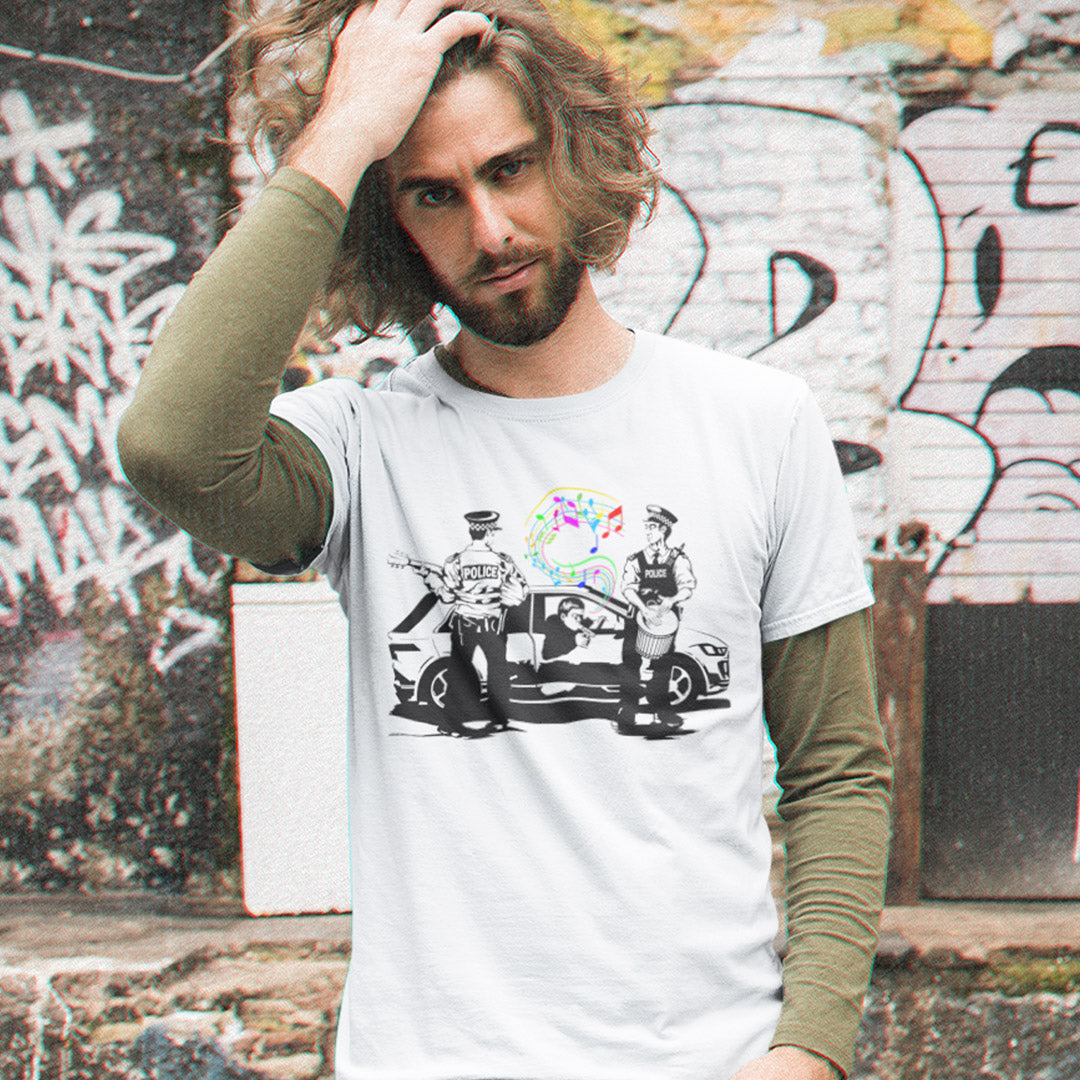 A man with long hair and a beard wearing a white graphic t-shirt over an olive green long-sleeve shirt, standing in front of a graffiti-covered wall.