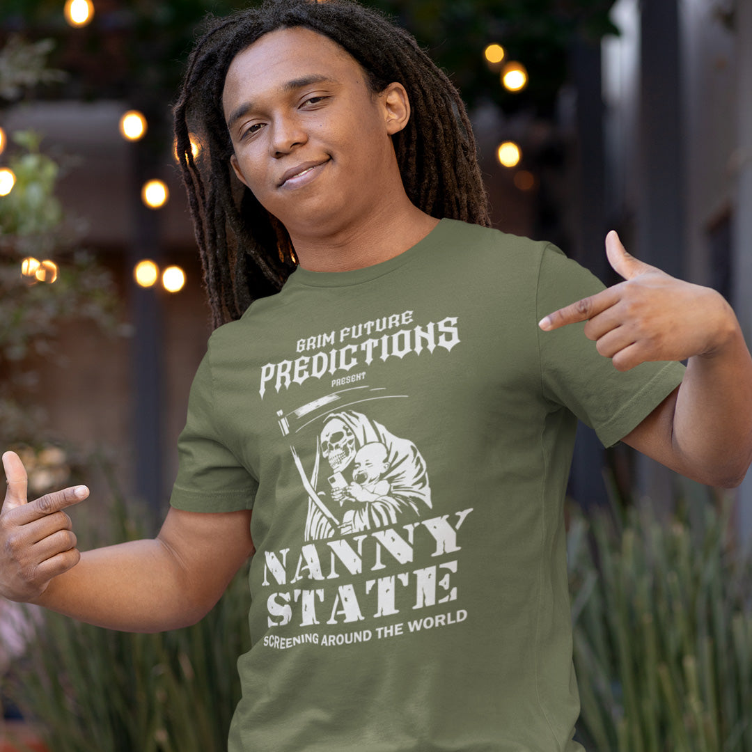 A man wearing an olive green graphic t-shirt with a white print that reads 'GRIM FUTURE PREDICTIONS PRESENTS NANNY STATE SCREENING AROUND THE WORLD' alongside an illustration of a nurse and a baby. He is posing outdoors with blurred lights and plants in the background.