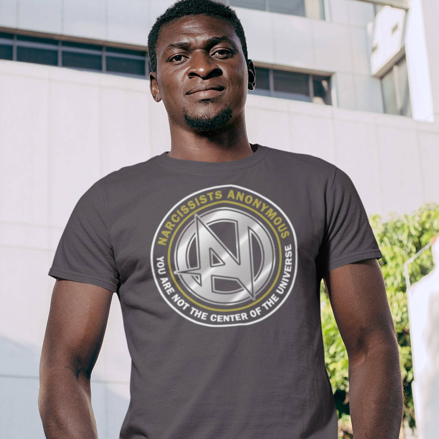 Man wearing an anthracite t-shirt with a circular logo and text outdoors. A satirical counter culture t shirt design suggesting narcissism is an illness in need of treatment. Wouldn't it be good if there was a place narcissists could go to get therapy and support for their disease. Made from 100% organic ring-spun cotton, this emotional intelligence t-shirt is unisex, high-quality, super comfy, and best of all, eco-friendly.