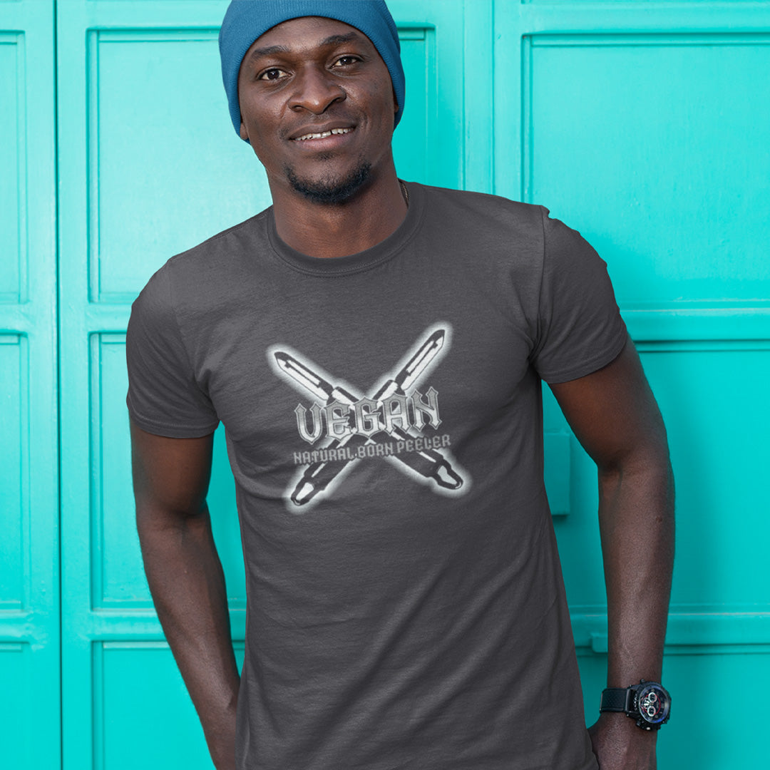 A smiling man standing in front of blue shutter doors, wearing a blue hat and an anthracite grey short sleeved crew neck t-shirt with a graphic of two crossed vegetable peelers and the text "VEGAN, NATURAL BORN PEELER" printed over the top.
