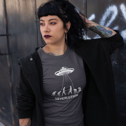 A woman with dark hair and tattoos wearing a dark gray t-shirt with a white UFO and evolution graphic, styled with a black open-front cardigan, posing against a dark textured wall.
