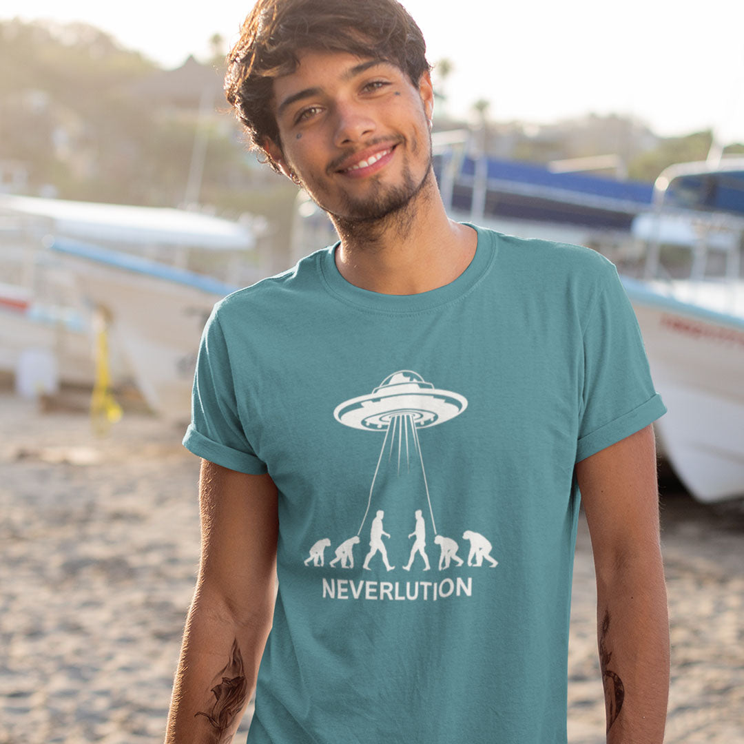 A man wearing a teal t-shirt with a white graphic of a UFO and the word 'NEVERLUTION' printed on the chest, standing on a sandy beach with boats in the background.