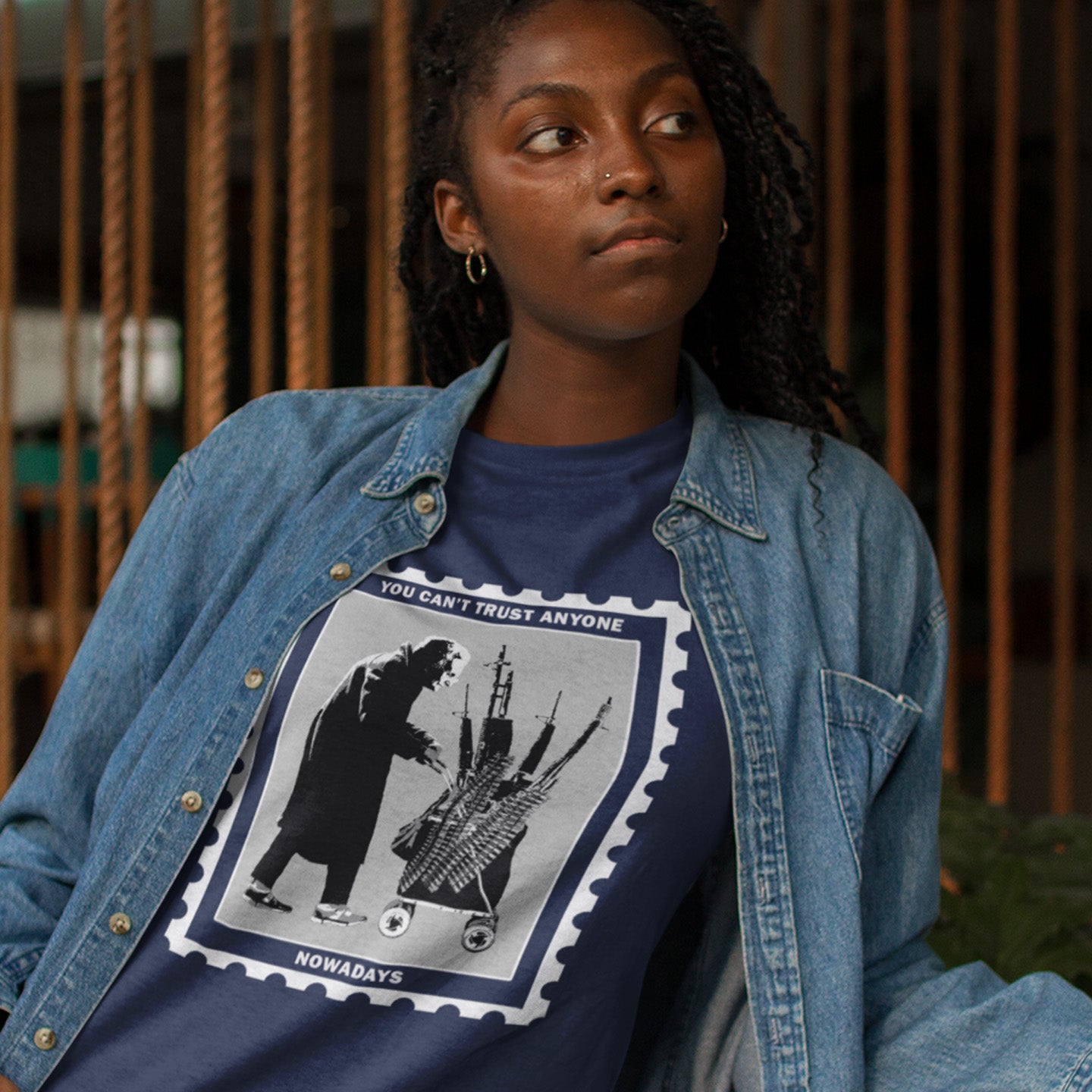 Person wearing a blue denim jacket over a graphic t-shirt with text and images. A Banksy style street art stencil t-shirt displaying a humorous view of how careful you have to be living in a culture of fear when you come from a more trusting generation. Made from 100% organic ring-spun cotton, this street art t-shirt is unisex, high-quality, super comfy, and best of all, eco-friendly.