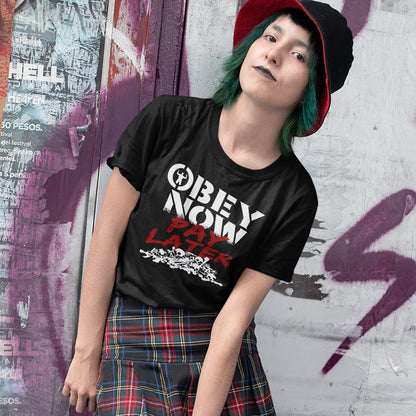 Person wearing a black t-shirt with 'OBEY NOW PAY LATER' text, standing against a graffiti-covered wall. This broken society t shirt highlights our need to act against corruption. The longer we ignore and comply to a broken system now the more we will pay for it later. Made from 100% organic ring-spun cotton, this anti government t shirt is unisex, high-quality, super comfy, and best of all, eco-friendly.