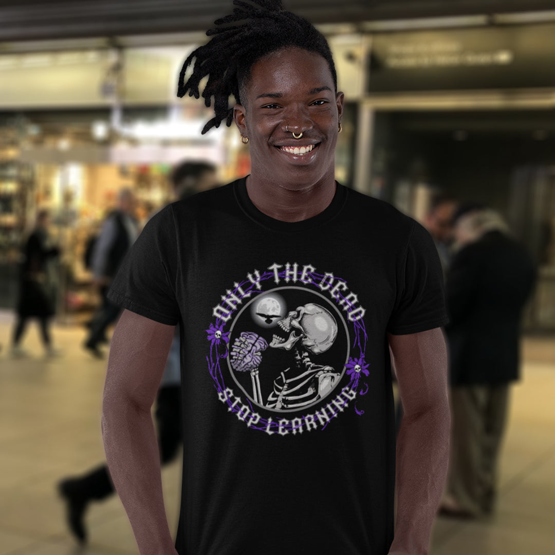 Person wearing a black t-shirt with a graphic design and text, standing in a blurred indoor setting. These thought provoking t shirts highlight our laziness in learning. Education is a life long experience that evolves the species. When schooling ends the real learning starts. Learning is for life, unless your brain has died on you. Made from 100% organic ring-spun cotton, this counter culture t shirt is unisex, high quality, super comfy, and best of all, eco-friendly.