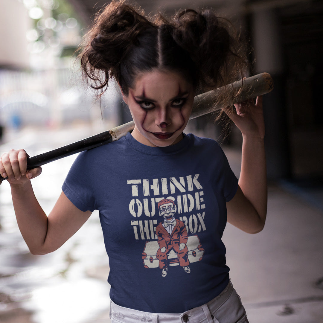 A woman with clown makeup and a baseball bat, wearing a French navy  t-shirt featuring a graphic print on the front. The graphic displays the text 'THINK OUTSIDE THE BOX' in a distressed beige font above an illustration of a ventriloquist dummy.