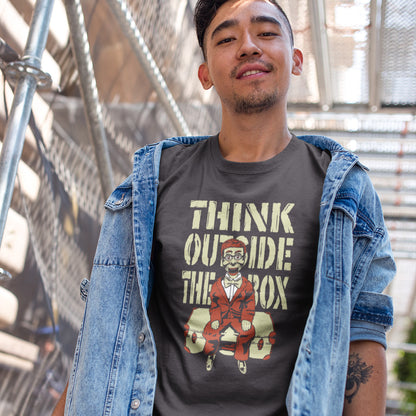 A man standing in the street, wearing an anthracite  t-shirt featuring a graphic print on the front. The graphic displays the text 'THINK OUTSIDE THE BOX' in a distressed beige font above an illustration of a ventriloquist dummy.