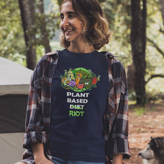 A woman wearing a navy blue graphic t-shirt with a cartoon vegetable illustration and the text 'PLANT BASED DIET RIOT' in white and green. She is wearing an open brown and red plaid flannel shirt over her shoulders, standing outdoors in a wooded campsite setting with a tent and a dog visible.