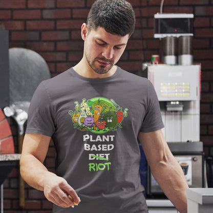 A man wearing an anthracite grey t-shirt with a colourful cartoon graphic of plants and the text 'PLANT BASED DIET RIOT' in a kitchen setting with a brick wall background.