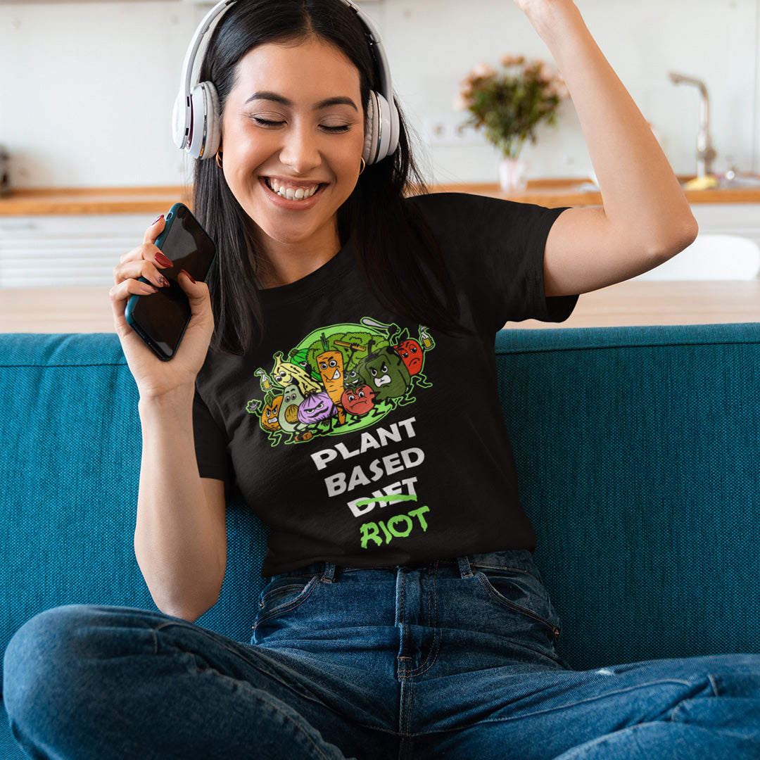 Woman enjoying music, wearing headphones, sitting on a couch, wearing a black t-shirt featuring a graphic print of a green chili pepper and a red chilli character with the text 'SPICY VEGAN' underneath.