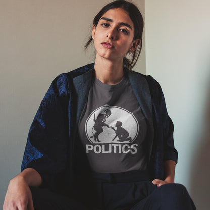 A woman sitting in a corner, wearing an anthracite t-shirt with a white graphic print on the chest. The graphic features a circular silhouette illustration of a standing figure reaching toward a kneeling figure, with the word 'POLITICS' printed in bold white capital letters underneath.