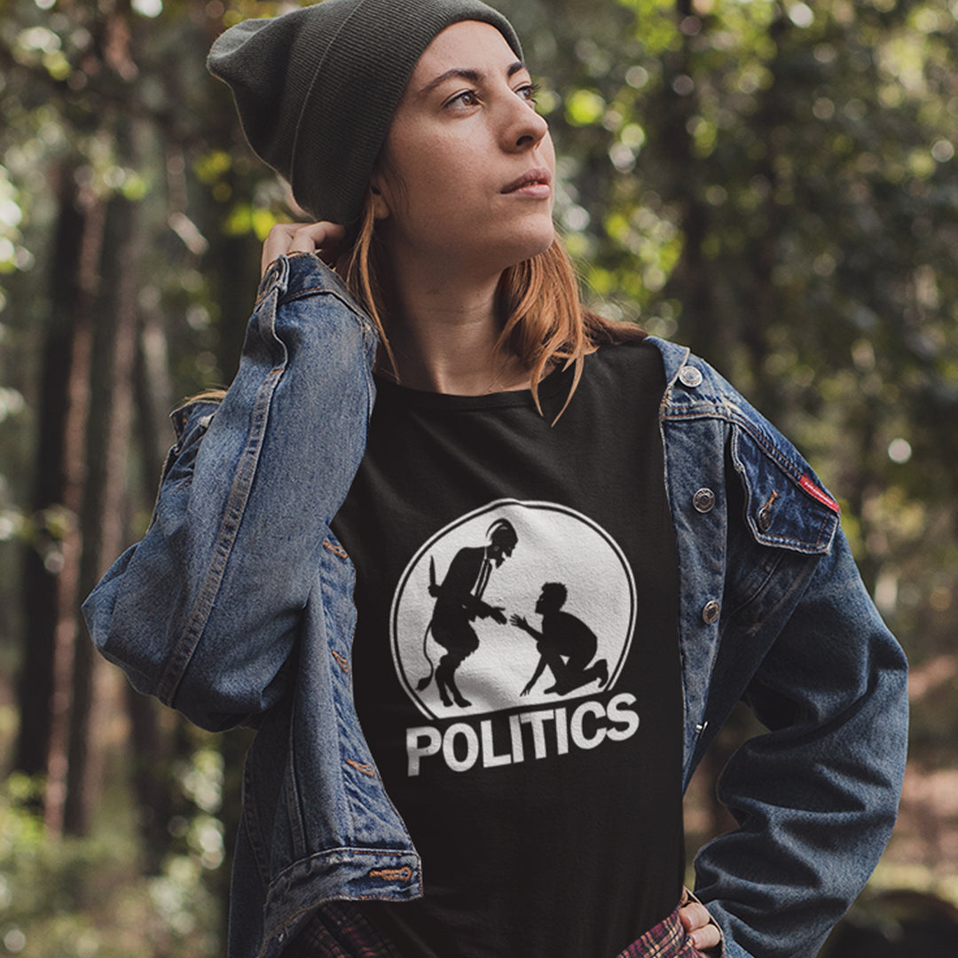A woman in a green hat, standing outdoors, wearing a black t-shirt with a white graphic print on the chest. The graphic features a circular silhouette illustration of a standing figure reaching toward a kneeling figure, with the word 'POLITICS' printed in bold white capital letters underneath.