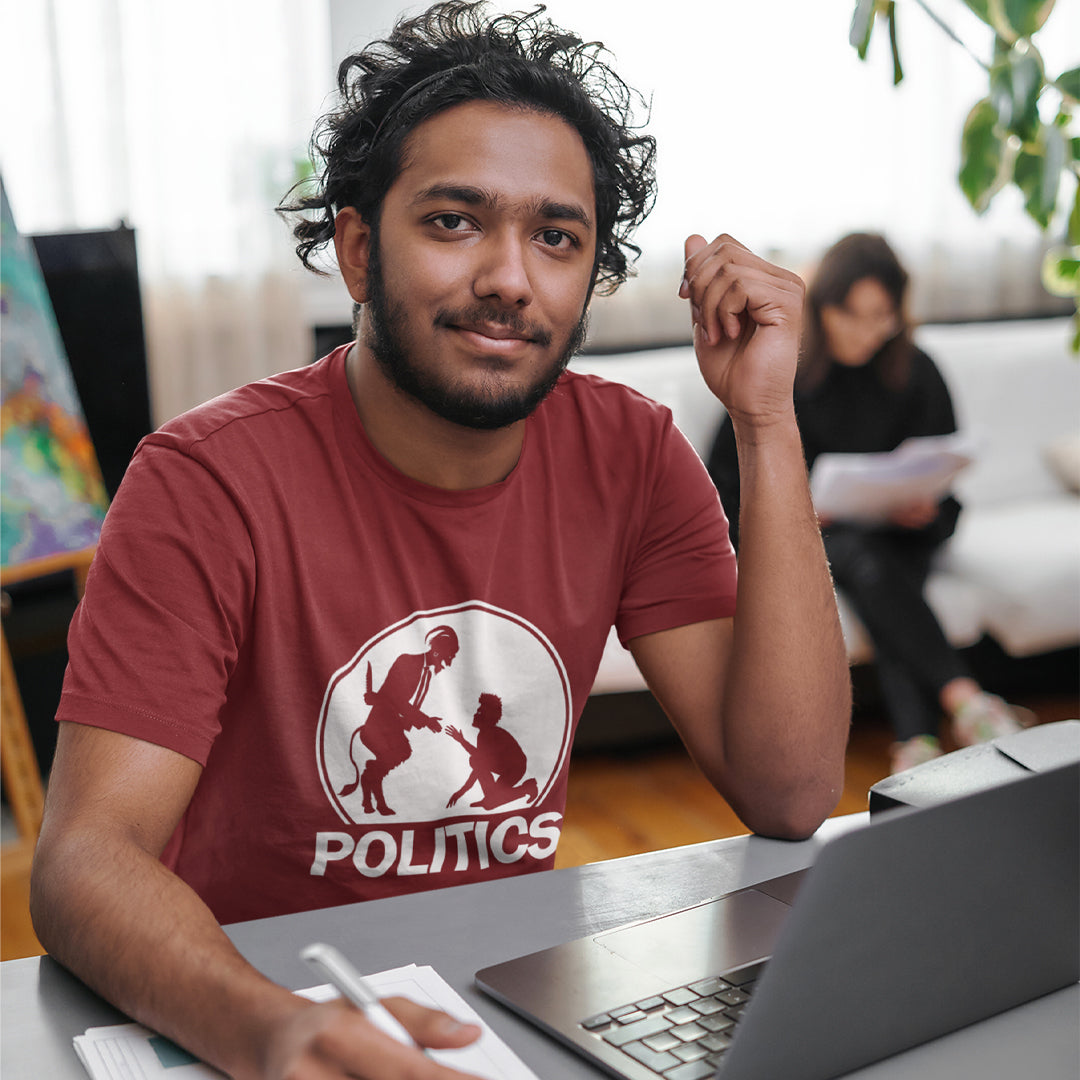 A man sitting at a desk with a laptop, wearing a burgundy t-shirt with a white graphic print on the chest. The graphic features a circular silhouette illustration of a standing figure reaching toward a kneeling figure, with the word 'POLITICS' printed in bold white capital letters underneath.