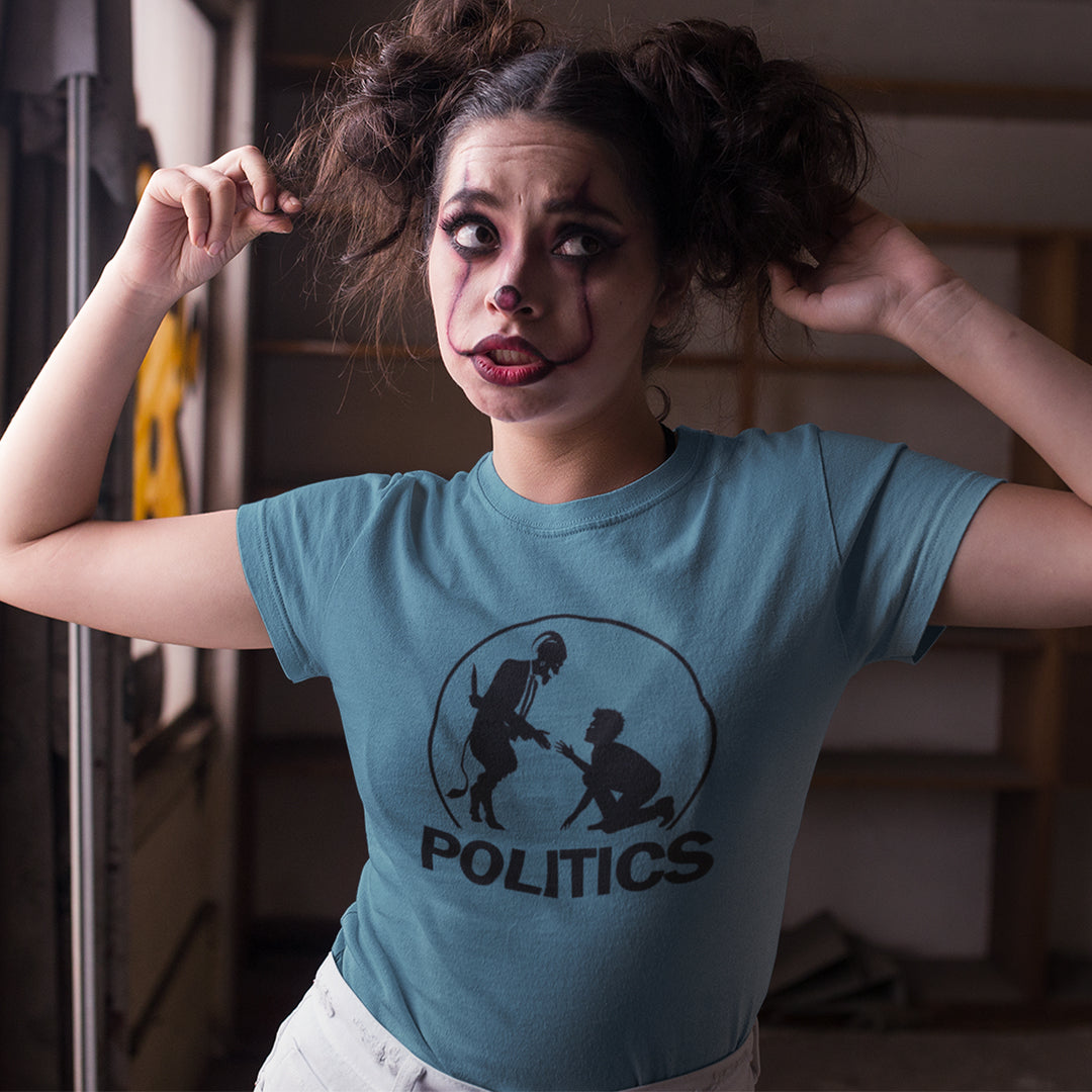 A woman with clown makeup wearing a stargazer blue t-shirt  with a black graphic print on the chest. The graphic features a circular silhouette illustration of a standing figure reaching toward a kneeling figure, with the word 'POLITICS' printed in bold white capital letters underneath.