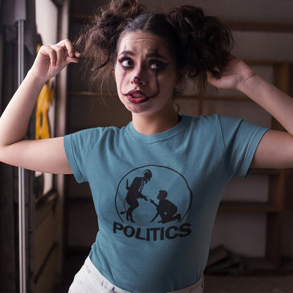 A woman with clown makeup wearing a stargazer blue t-shirt  with a black graphic print on the chest. The graphic features a circular silhouette illustration of a standing figure reaching toward a kneeling figure, with the word 'POLITICS' printed in bold white capital letters underneath.