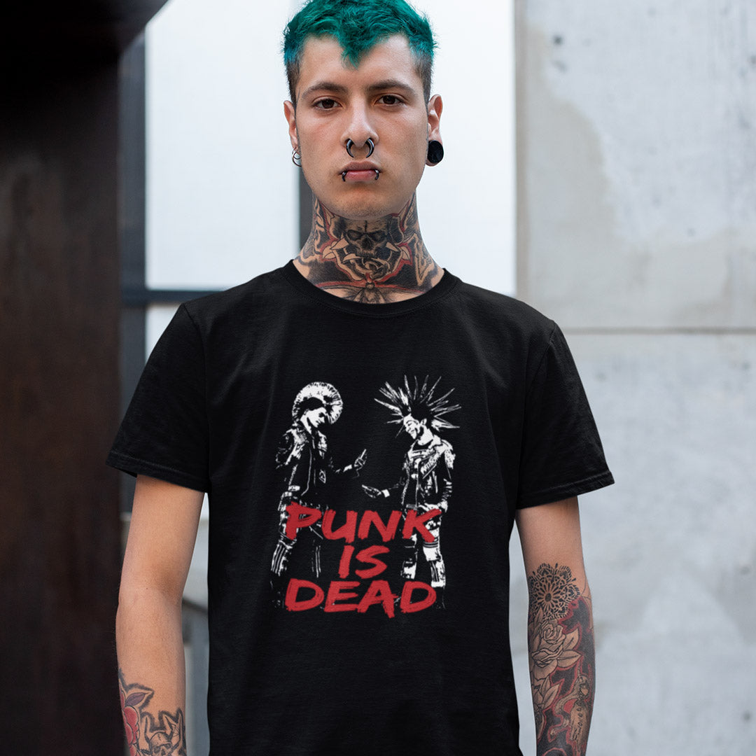A man with green hair, tattoos, and nose piercings wearing a black graphic t-shirt with the text 'PUNK IS DEAD' in red and white, standing against a concrete wall.