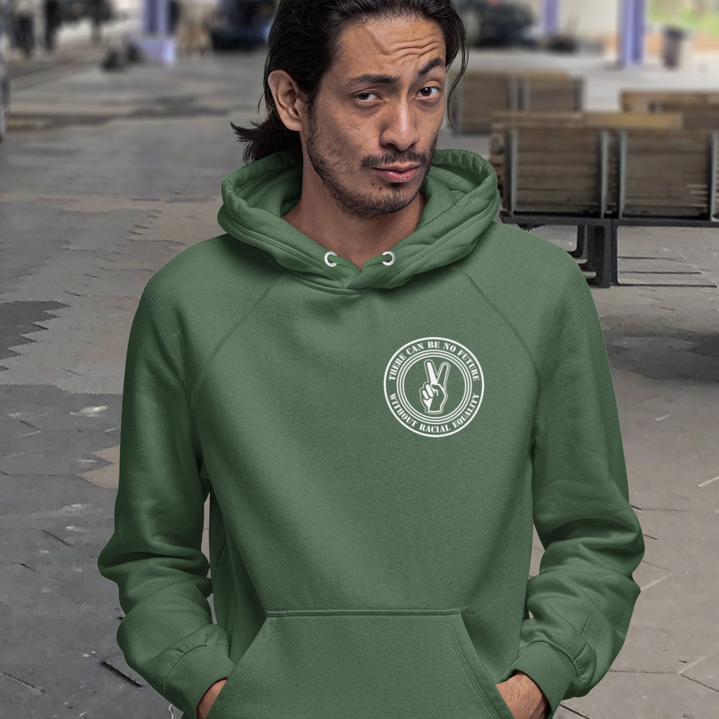 Person wearing a green hoodie with a logo on a street background. An anti-racism hoodie for Peace without prejudice There can be no future without racial equality. Comfortable and practical. It's extra soft and has a convenient front pouch pocket. Made from organic cotton and recycled polyester, this anti racism hoodie is a great eco-friendly choice.