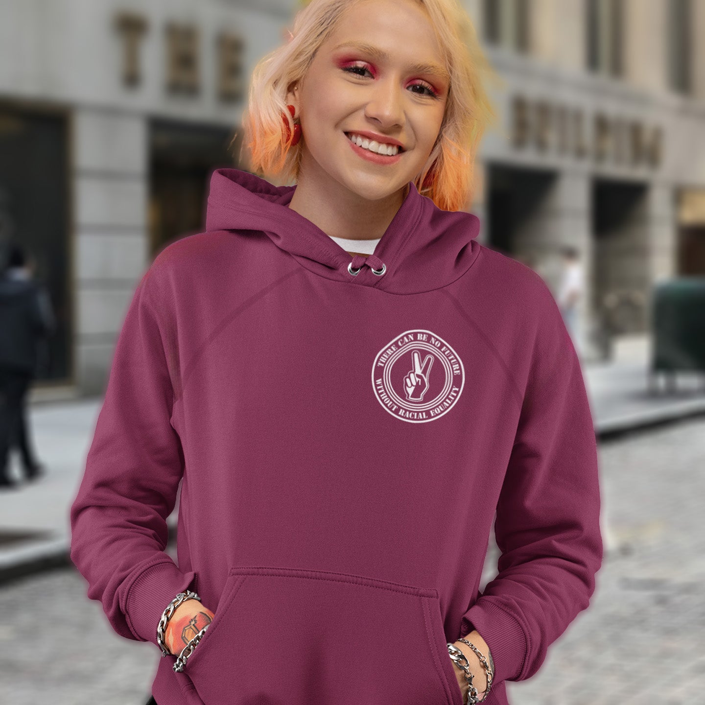 Person wearing a maroon hoodie with a logo, standing on a city street. An anti-racism hoodie for Peace without prejudice There can be no future without racial equality. Comfortable and practical. It's extra soft and has a convenient front pouch pocket. Made from organic cotton and recycled polyester, this anti racism hoodie is a great eco-friendly choice.