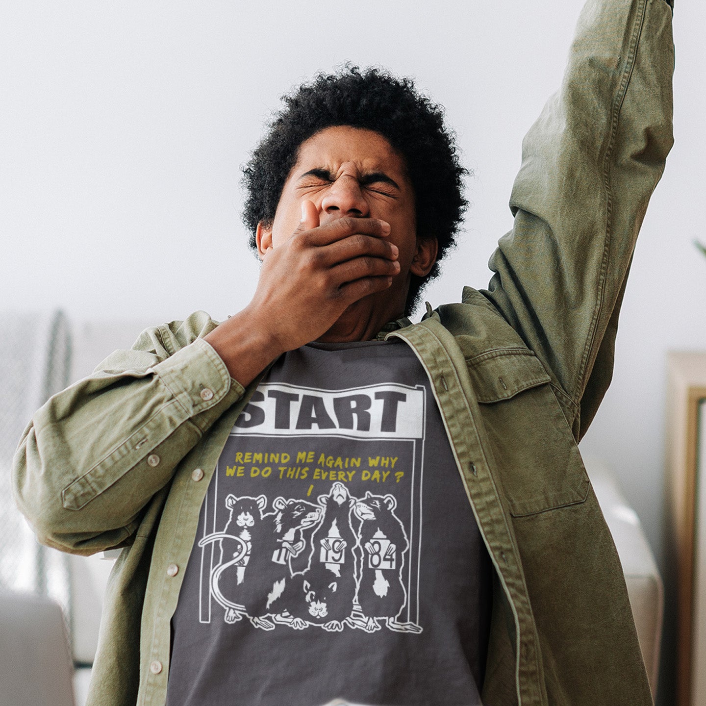 Person wearing a t-shirt with a graphic design and text, indoors. This stencil graffiti t shirt represents the monotony of everyday working life. A unique satirical design for those times when life starts to feel a little repetitive, unnecessary, and questionable. Made from 100% organic ring-spun cotton, this urban art t shirt is unisex, high-quality, super comfy, and best of all, eco-friendly.