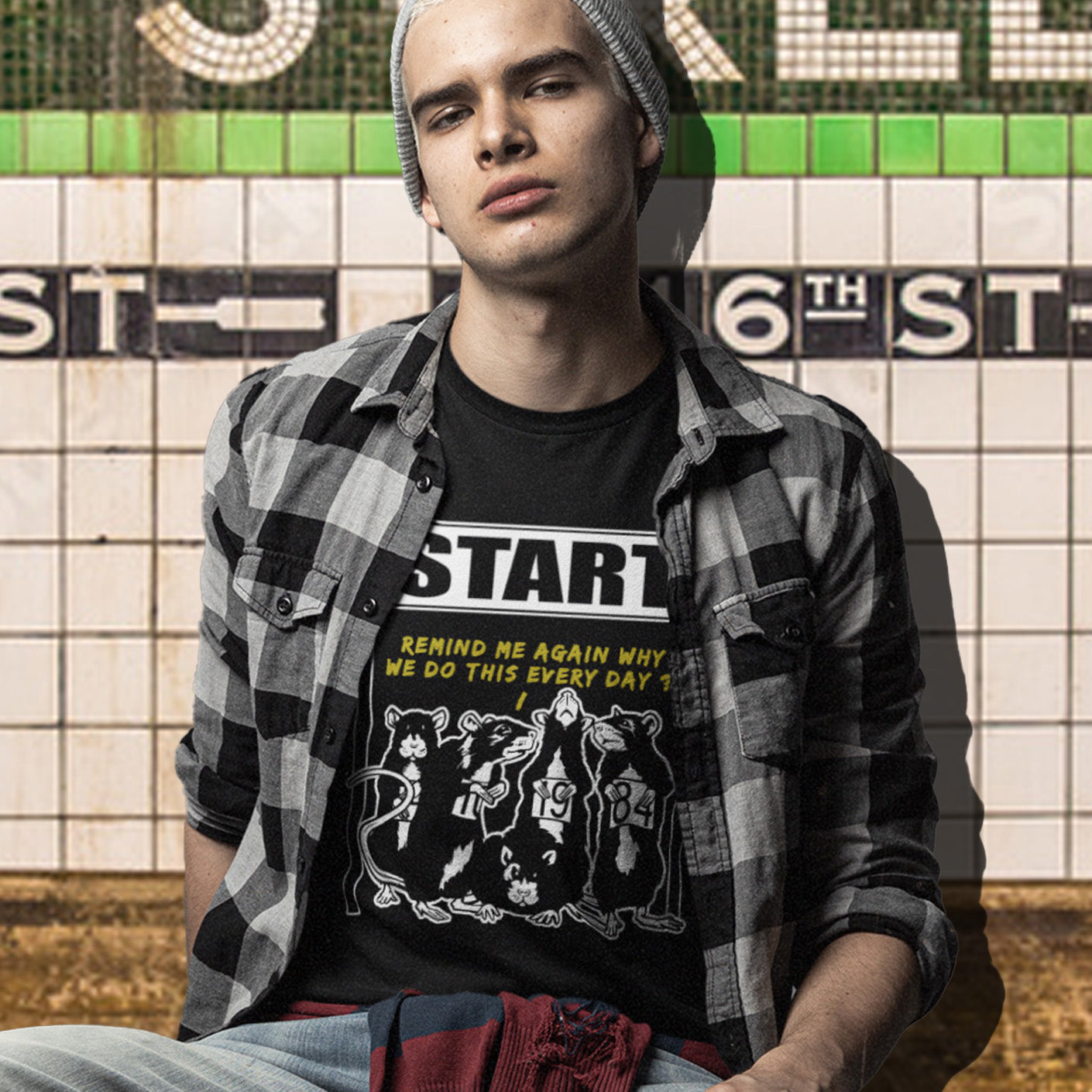Person wearing a black t-shirt with graphic design and text, sitting against a tiled wall. This stencil graffiti t shirt represents the monotony of everyday working life. A unique satirical design for those times when life starts to feel a little repetitive, unnecessary, and questionable. Made from 100% organic ring-spun cotton, this urban art t shirt is unisex, high-quality, super comfy, and best of all, eco-friendly.