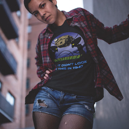 Person wearing a black t-shirt with graphic design and text, standing against an urban background. A funny alien t shirt for lovers of conspiracy theory. Imagine the aliens from the 1947 Roswell incident coming back in the future to see what we've done with the place. Made from 100% organic ring-spun cotton, this funny alien t shirt is unisex, high-quality, super comfy, and best of all, eco-friendly.