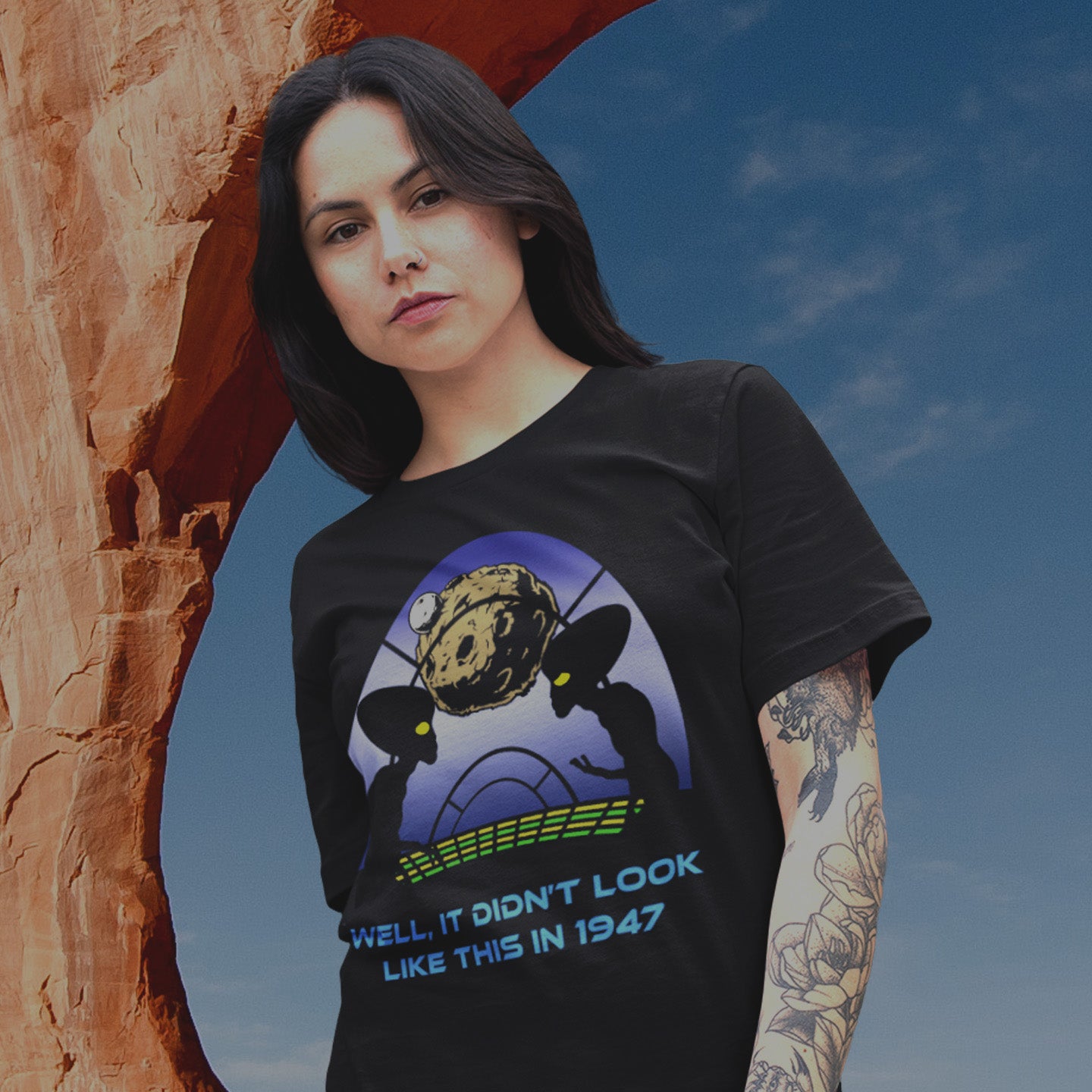 Woman wearing a black t-shirt with a graphic design and text, standing in front of a natural rock formation. A funny alien t-shirt for lovers of conspiracy theory. Imagine the aliens from the 1947 Roswell incident coming back in the future to see what we've done with the place. Made from 100% organic ring-spun cotton, this witty alien t-shirt is unisex, high-quality, super comfy, and best of all, eco-friendly.