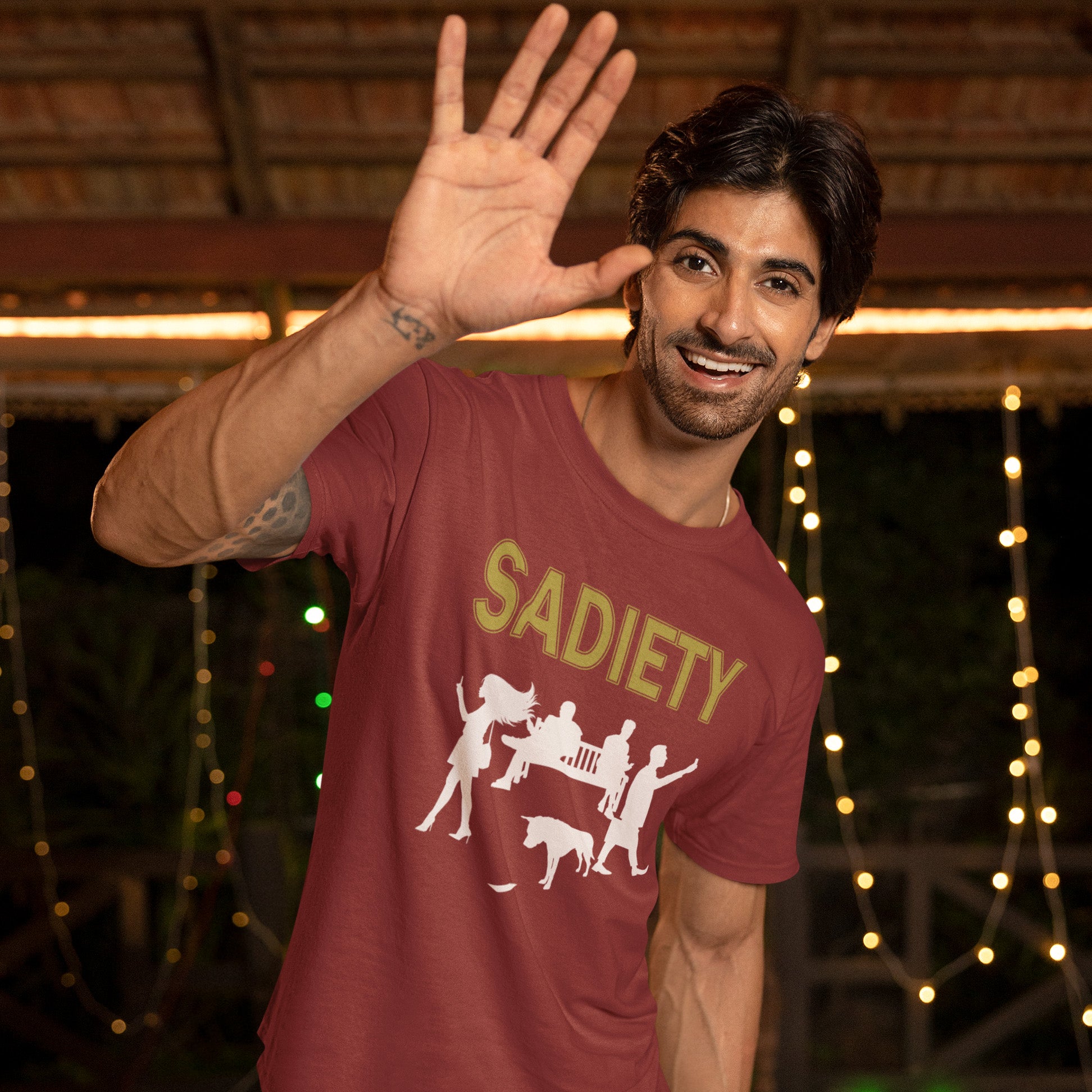 Man wearing a burgundy t-shirt with 'SADIETY' design, waving at the camera with string lights in the background. An anti mobile phone abuse t shirt design promoting social awareness about the effects of the mobile phone on society and sociability. The mobile phone has made people in close proximity seem miles apart. It's not a society, it's sadiety. Made from 100% organic ring-spun cotton, this stencil graffiti t shirt is unisex, high-quality, super comfy, and best of all, eco-friendly.