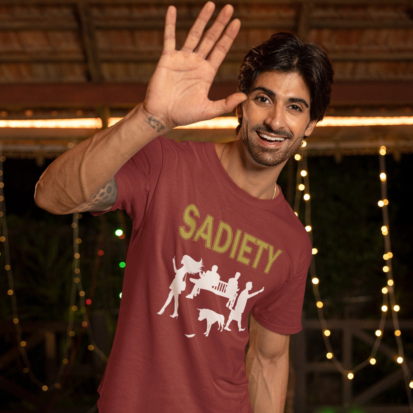 Man wearing a burgundy t-shirt with 'SADIETY' design, waving at the camera with string lights in the background. A counterculture t-shirt design promoting social awareness about the effects of the mobile phone on society and sociability. The mobile phone has made people in close proximity seem miles apart. It's not a society, it's sadiety. Made from 100% organic ring-spun cotton, this subculture t-shirt is unisex, high-quality, super comfy, and best of all, eco-friendly.